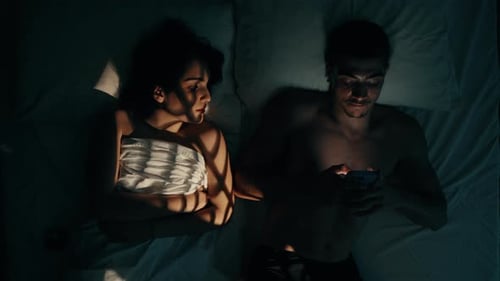 Man Using Smartphone in Bed While Woman Tries to Sleep