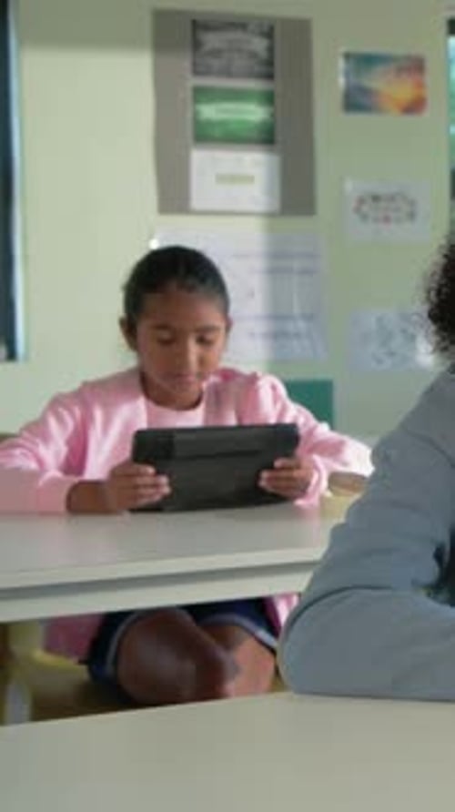 Vertical video: Sitting girl in pink top doing work on tablet in class, glancing as peer moving in