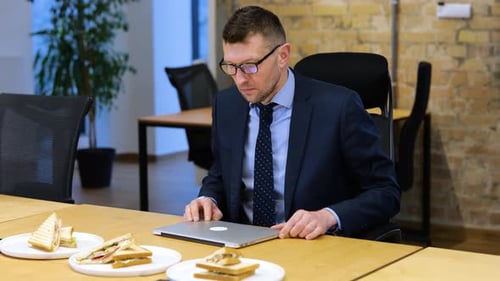 Kaukasischer Mann arbeitet an seinem Laptop, dann hört er auf zu arbeiten und fängt an, drei Sandwiches zu essen