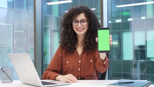 Smiling businesswoman showing mobile phone with green screen sitting at workplace in business office