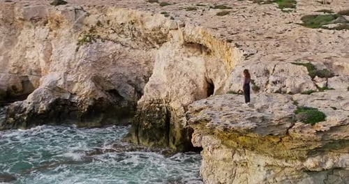 Aerial View of a Woman Standing on the Edge of a Rocky Cliff on the Seashore