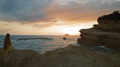 Young girl walks on rocky cliff at seaside watching sunset over the ocean in Kefalonia