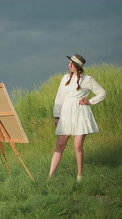 Woman in White Dress and Hat Posing By Canvas in Grassy Field