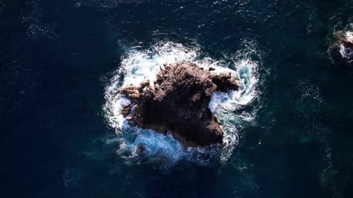 Aerial top down view of waves smashing a cliff in middle of blue ocean