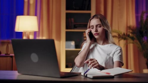 A Woman is Working Late at Her Home Office Using a Laptop and Talking on the Phone