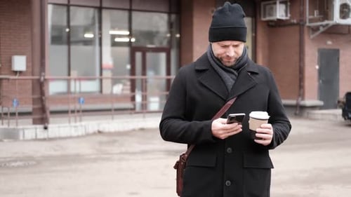 Handsome Man Wearing Black Coat and Brown Leather Bag Walking Down on the City Drinking Coffee Paper