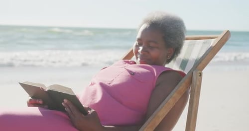 Happy senior african american woman sitting on deck chair and reading book at beach, in slow motion