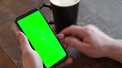 Mann benutzt Handy mit leerem grünem Bildschirm im Café auf einem rustikalen Vintage-Holztisch