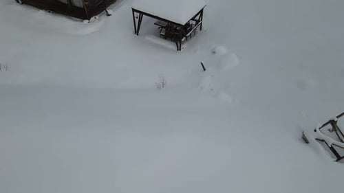 Drone flies over fence, mountain cottage towards pine forest, everything covered in thick, white sno