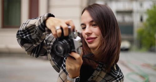Happy Brunette Girl in a Plaid Shirt Holding a Camera and Taking a Photo Smiling Widely in the City