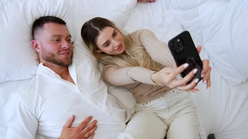 Das kaukasische Paar liegt zusammen auf dem Bett. Attraktive Frau macht Selfie von sich und ihrem Mann
.