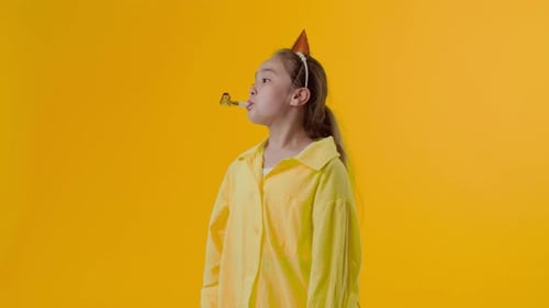 Young Asian Girl in the Paper Cap Blowing a Party Horn and Looking at the Camera with an Unhappy