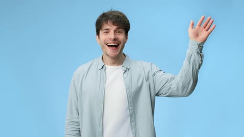 Man Waves With a Friendly Smile Against a Light Blue Background in Bright Lighting While Wearing a