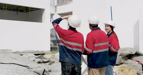 Männlicher Bauunternehmer zeigt auf die Außenwand, während die asiatische Ingenieurin den Gerüstbereich beobachtet,
