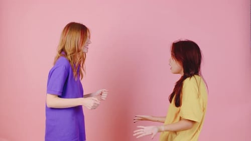 Two Close Friends Chat Animatedly Against a Pink Background Dressed in Colorful Outfits Sharing Joy