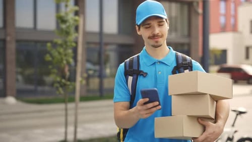 Smiling Courier Going Along Building Holding the Paper Boxes and Mobile Phone in Hand
