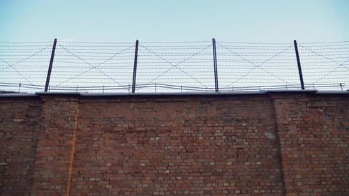 Low-angle view of jail prison dark red brick wall with a barbed wire fence on top with the cloudy sk