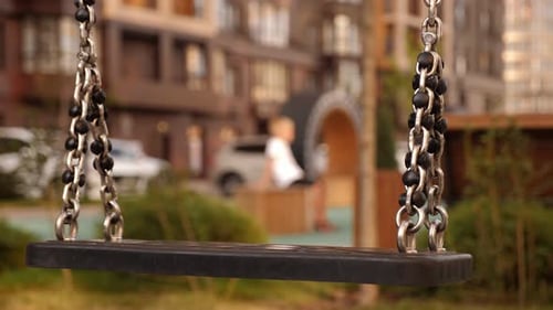 A Sad Boy Leaves the Playground Alone in the Foreground of a Swing on Chains