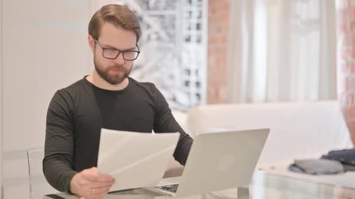 Man Reading Papers and Using Laptop at Desk