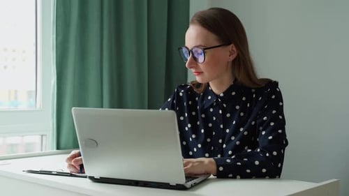 A Young Business Woman in Glasses and a Shirt is Working at a Laptop in a Professional Office