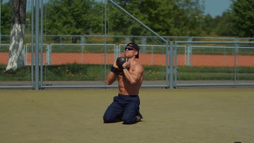 Bodybuilder in Black Cap and Sunglasses Sits on His Knees on Sports Ground and Lifts Kettlebell Up