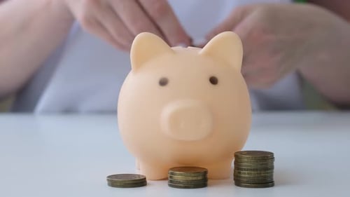 Female Hand Putting Coins Into Piggy Bank on White Table Saving Money Concept