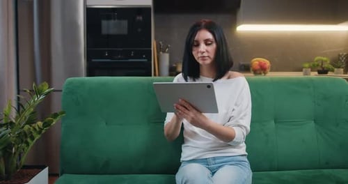 Joven mujer caucásica sonriente se sienta en el sofá de la cocina usando una tableta digital, las redes sociales, leyendo