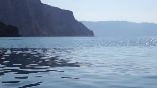 Sea Water Surface Camera Flies Over the Calm Azure Sea with Volcanic Rocky Shores on Background