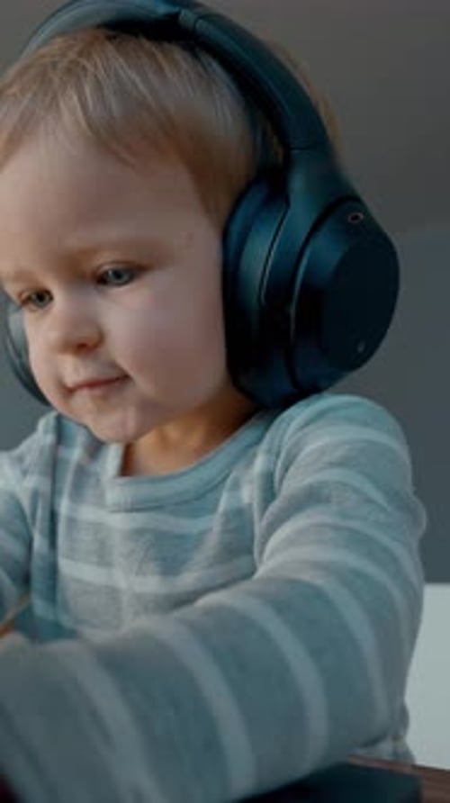 Vertical of Toddler Listens to Music in Headphones Typing on Laptop