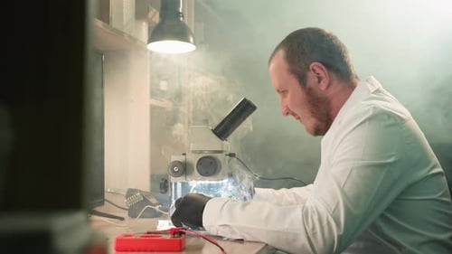 Technician Soldering Circuit Board Under Microscope in Electronic Repair Workshop