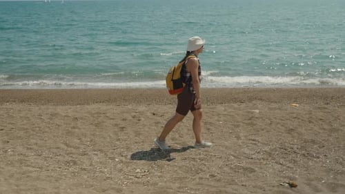 A Woman Walks Along the Beach, Wearing a Hat and Backpack, With Gentle Waves Crashing on the Shore B