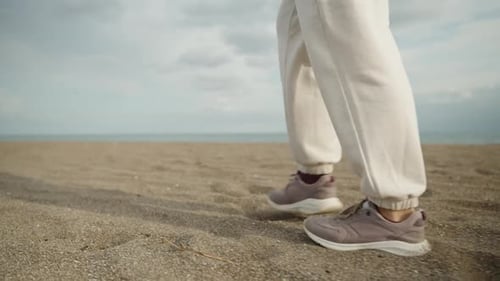 feet in purple sneakers and white pants are walking along a sandy beach along the sea, Cloudy, slow.