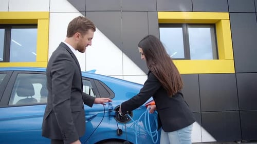 Businessman in a dealership shows a girl how to charge an electric car at a charging station