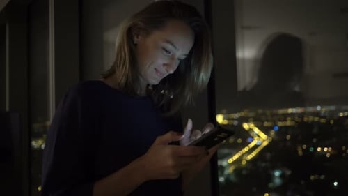 Smiling Woman Using Smartphone at Night Near Window