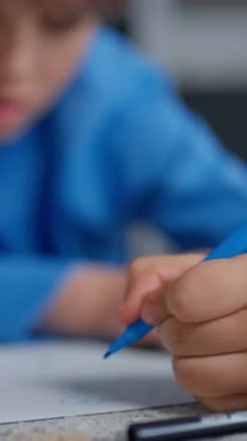 Closeup of a Young Child in a Blue Shirt Drawing with a Blue Pen on Paper Then Concentrating with a