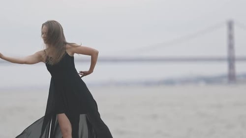 An Adult Woman in Black Dress Dancing on the Promenade in Cloudy Weather with a Bridge on the