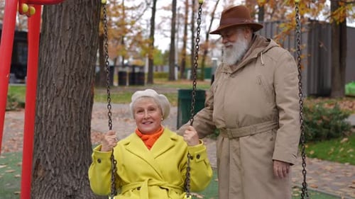 Smiling Senior Man Swinging Relaxed Woman on Swings at Children's Playground in Park