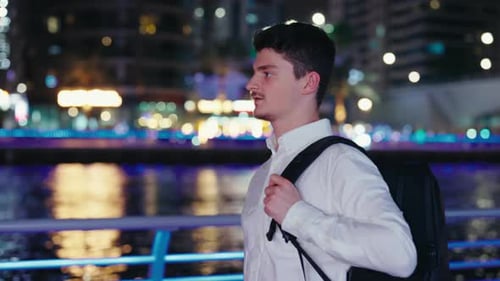 Side View of the Young Guy Walking on a Night Street Wearing a Backpack