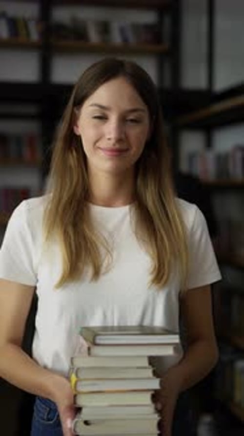 Student Girl Walks Through Library with Stack of Books in Hands Shelves with Book Front View Young
