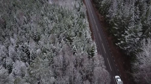 slow moving drone shot of a car driving up a road and driving through frosty forest and paved road.