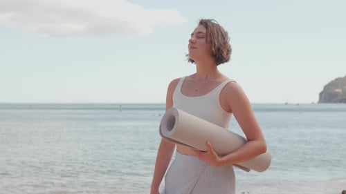 Smiling Woman with Rolled Yoga Mat Standing By Sea Under Bright Sky