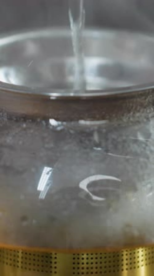 Close Up Pouring Hot Water Into Glass Kettle As Steam Rises with Golden Herbal Tea