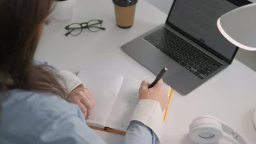 Woman Working at a Modern Desk with a Laptop Writing Notes in a Notebook While Studying Online