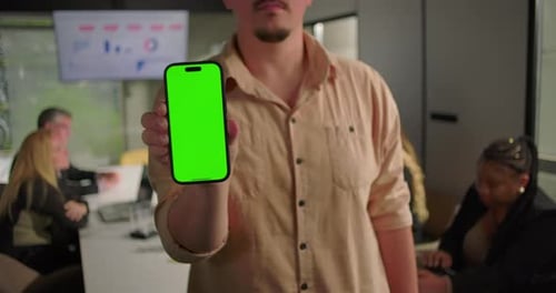 Young man holding smartphone with green screen in office, extending arm toward camera