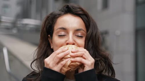 Young Businesswoman Standing Outdoors with Sandwich in Her Hands with Office Center on the