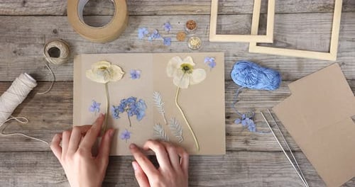 Woman putting dry flowers into notebook at wooden table, top view