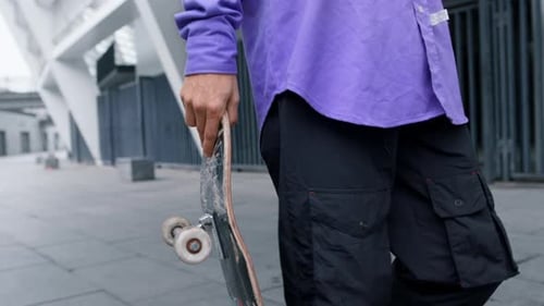 Young man holding skateboard walking through city street on bright summer day