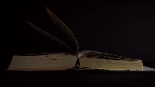 Slow motion, close up of an old, vintage, open book with pages turning on the wind, on a dark room w
