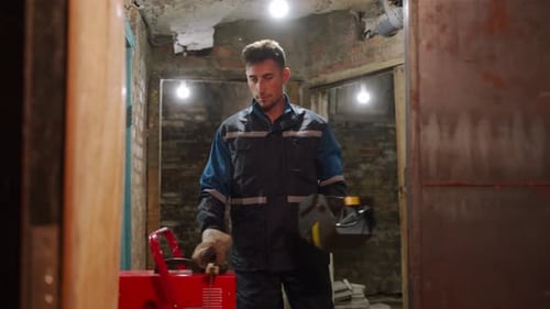 Professional Welder in Safety Suit Comes Up to Welding Machine Takes Electrode Holder and Puts on