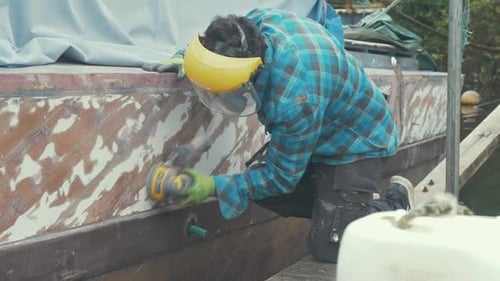 Um homem lixando o casco de um barco de madeira usando uma lixadeira orbital
